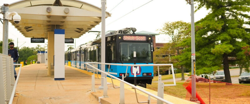A MetroLink train passes through UMSL North Station with construction fencing nearby.
